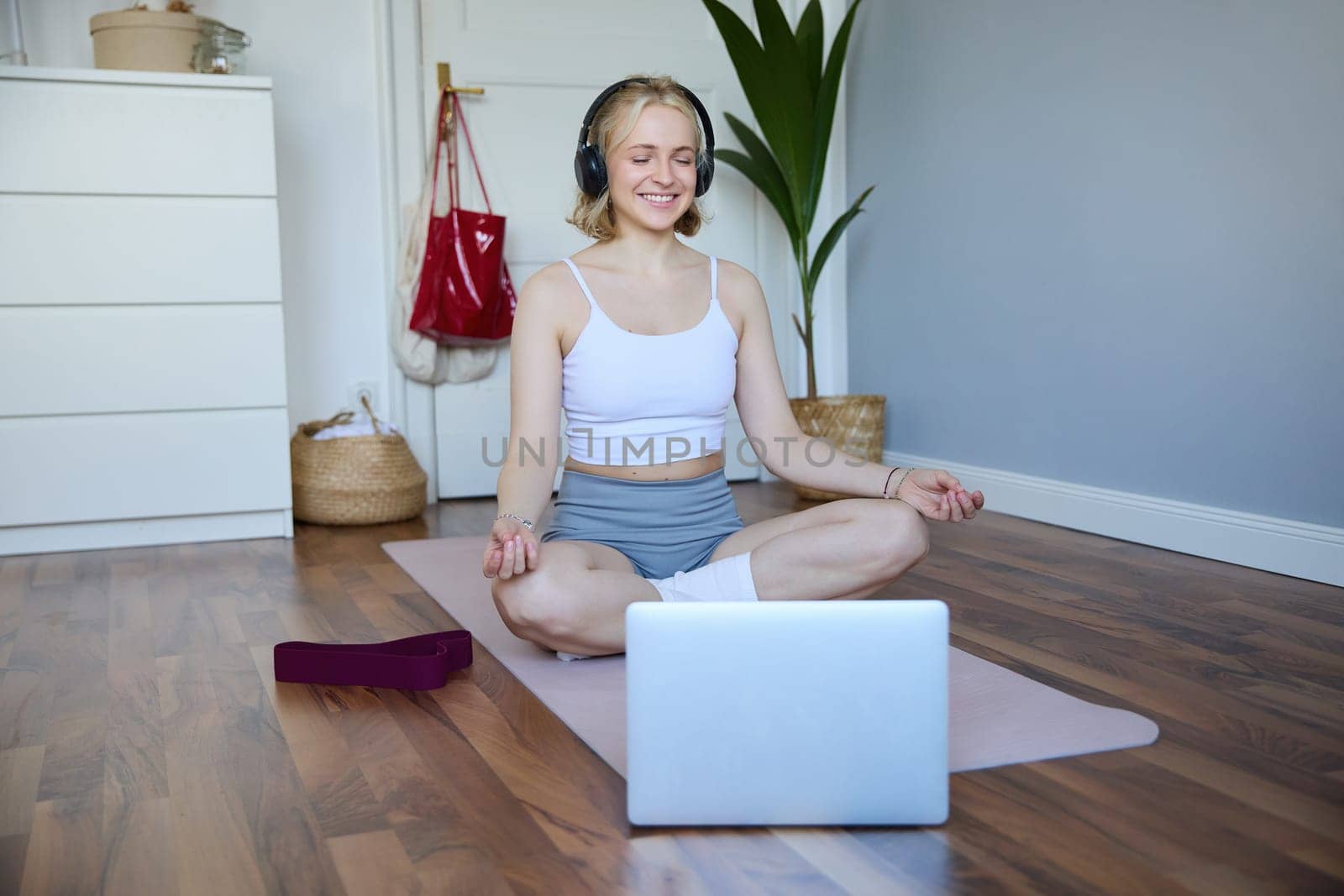 Portrait of young and relaxed woman using laptop and wireless headphones while meditating, practice yoga at home on rubber mat, sitting in asana.