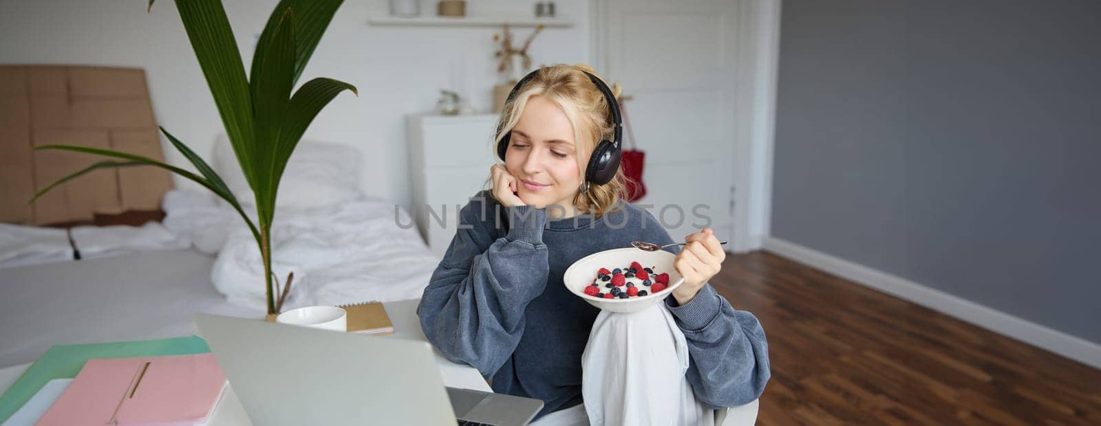 Image of happy woman sitting in a room, watching interesting tv show or movie on laptop, using screaming service, wearing headphones, eating dessert and drinking tea.