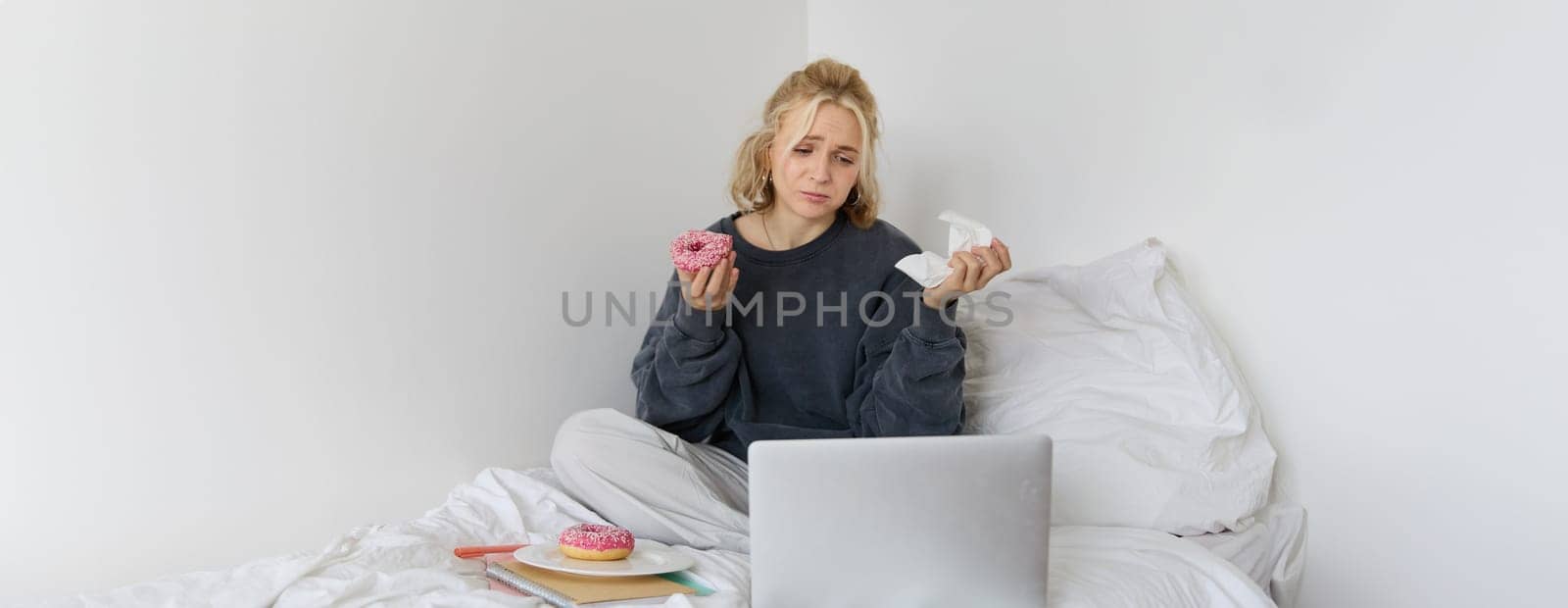Portrait of sad, crying young woman, staying at home, sitting in bed with doughnut and comfort food, looking at something upsetting on laptop screen by Benzoix