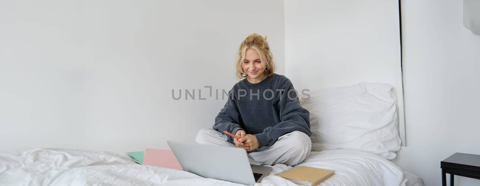 Portrait of happy blond woman, freelancer working from home, sitting on bed with laptop and notebooks. Student doing homework in bedroom, connects to online class via video chat by Benzoix