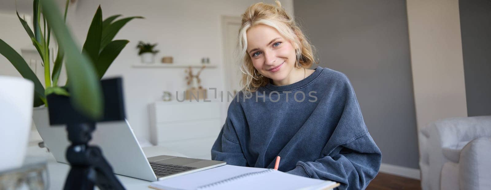 Portrait of beautiful smiling woman, student studying at home, remote education concept, sits in a room with laptop and writes down notes, uses her journal.