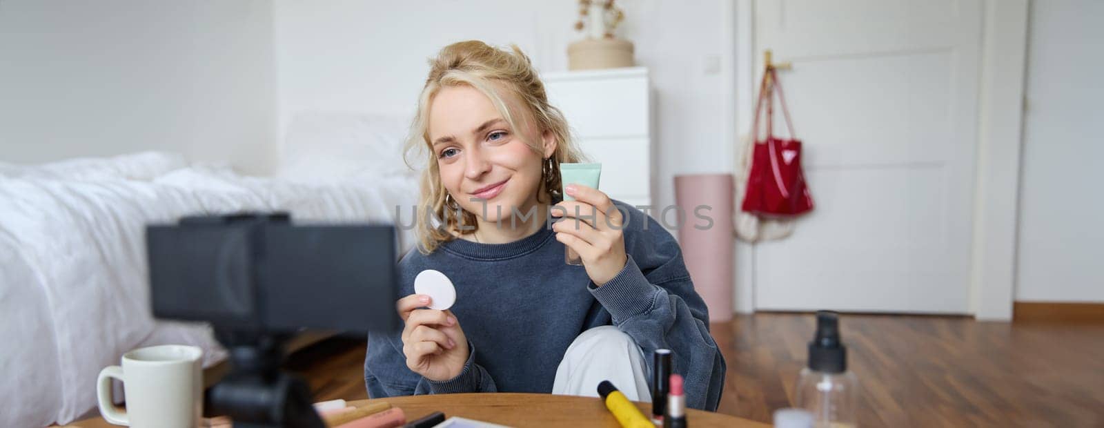 Portrait of young woman promoting beauty product, applies makeup in front of camera, recording video for her vlog, creating content for social media, sitting in a room on floor.