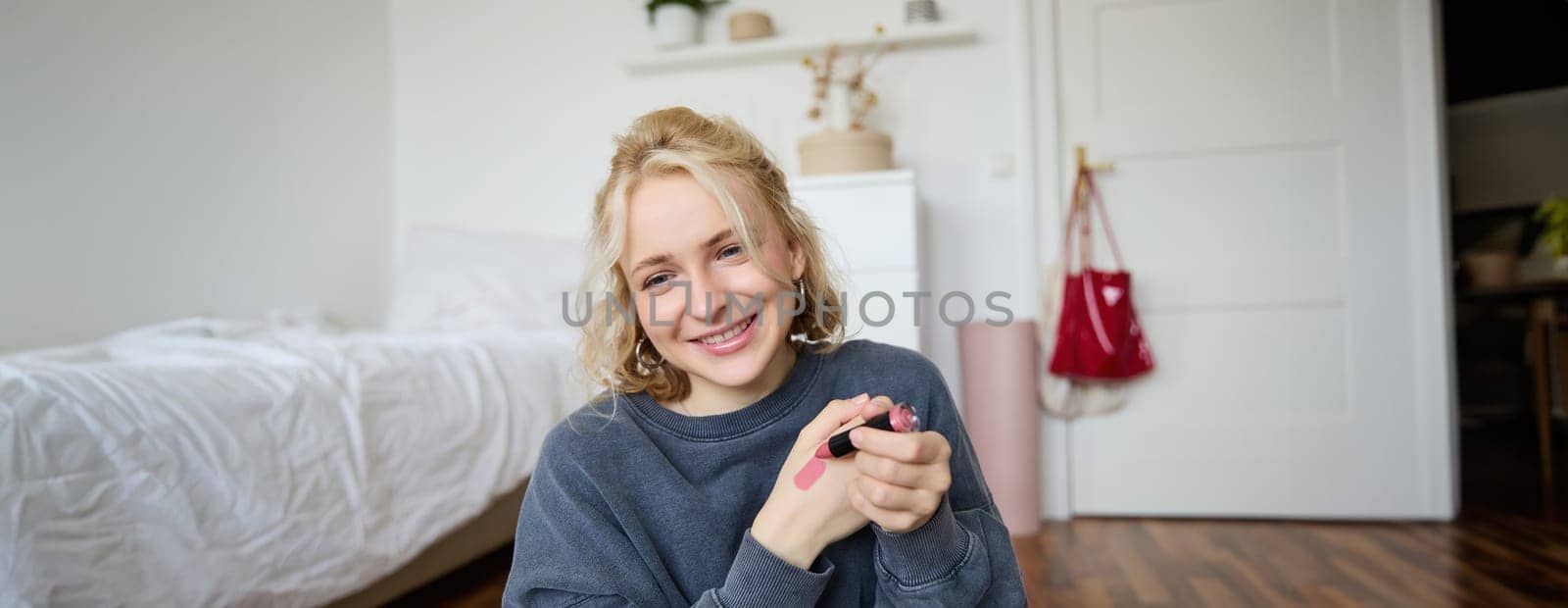 Portrait of beautiful young woman, content creator for social media, sitting in front of digital camera, recording video about makeup, showing lipstick swatches on her skin.