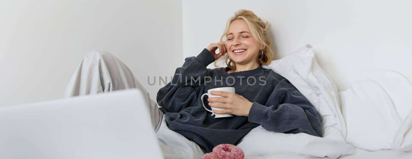 Portrait of happy woman enjoying day-off at home, lying in bed, watching tv show on laptop, looking at screen and smiling, drinking tea and eating in bedroom.