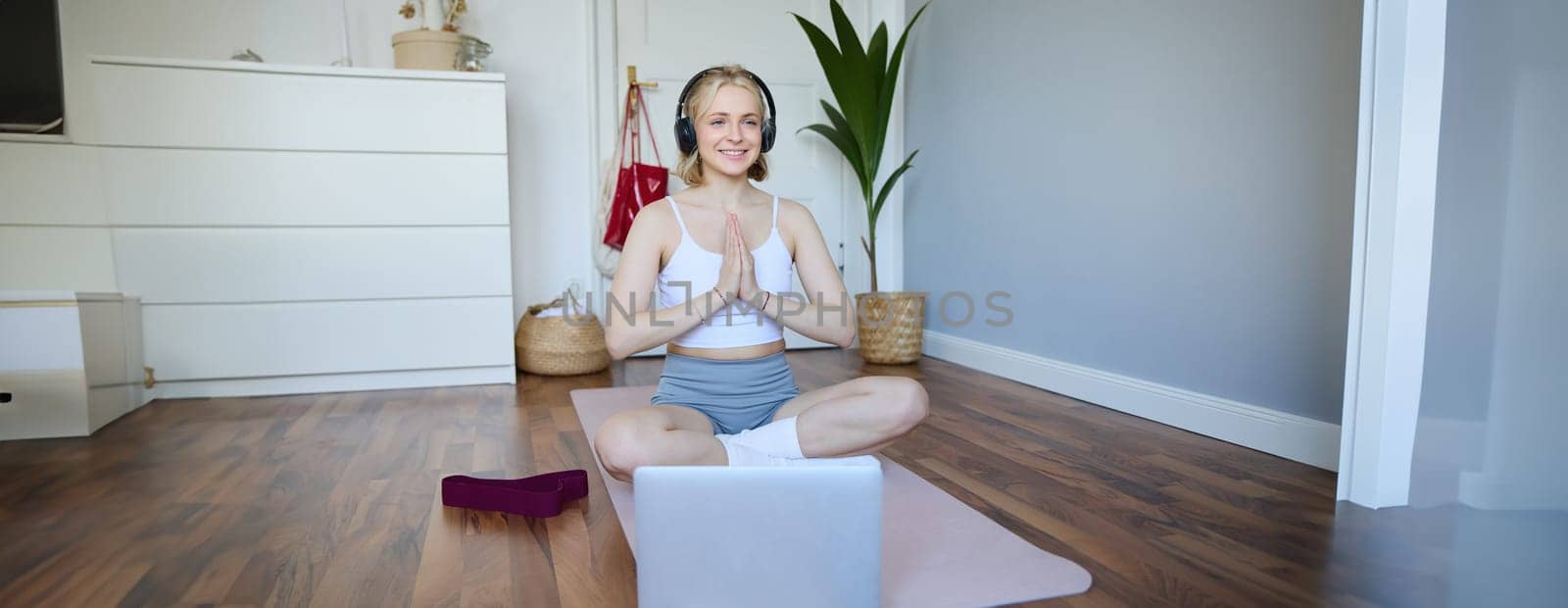 Portrait of blond young woman meditating, practise yoga, using yoga podcast to workout at home, sits on rubber mat.