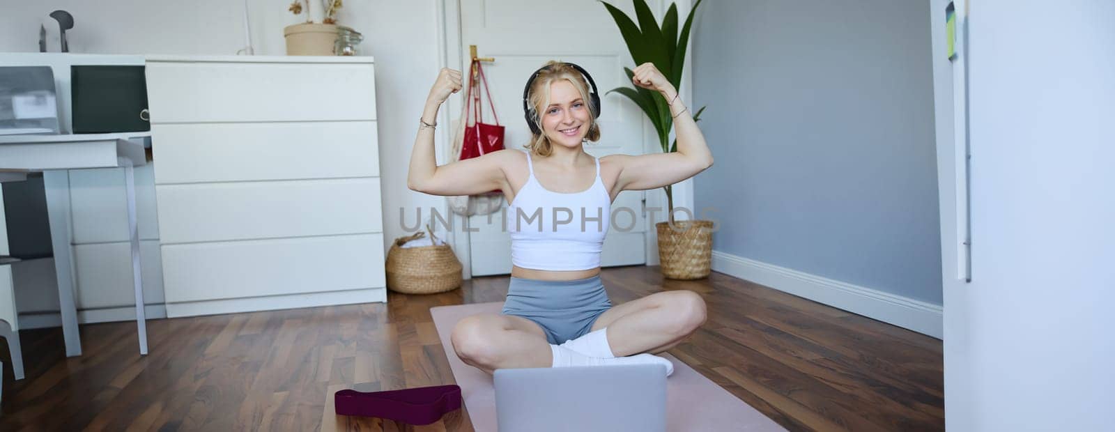 Portrait of young happy woman, fitness blogger in wireless headphones, working out at home, sits on rubber mat with laptop and wireless headphones, shows her muscles.