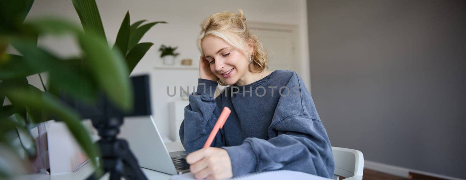 Cute smiling girl sits in a room, writes down notes, doing homework, records video of herself on digital camera, creates content for vlog, lifestyle blogger doing daily routine episode by Benzoix