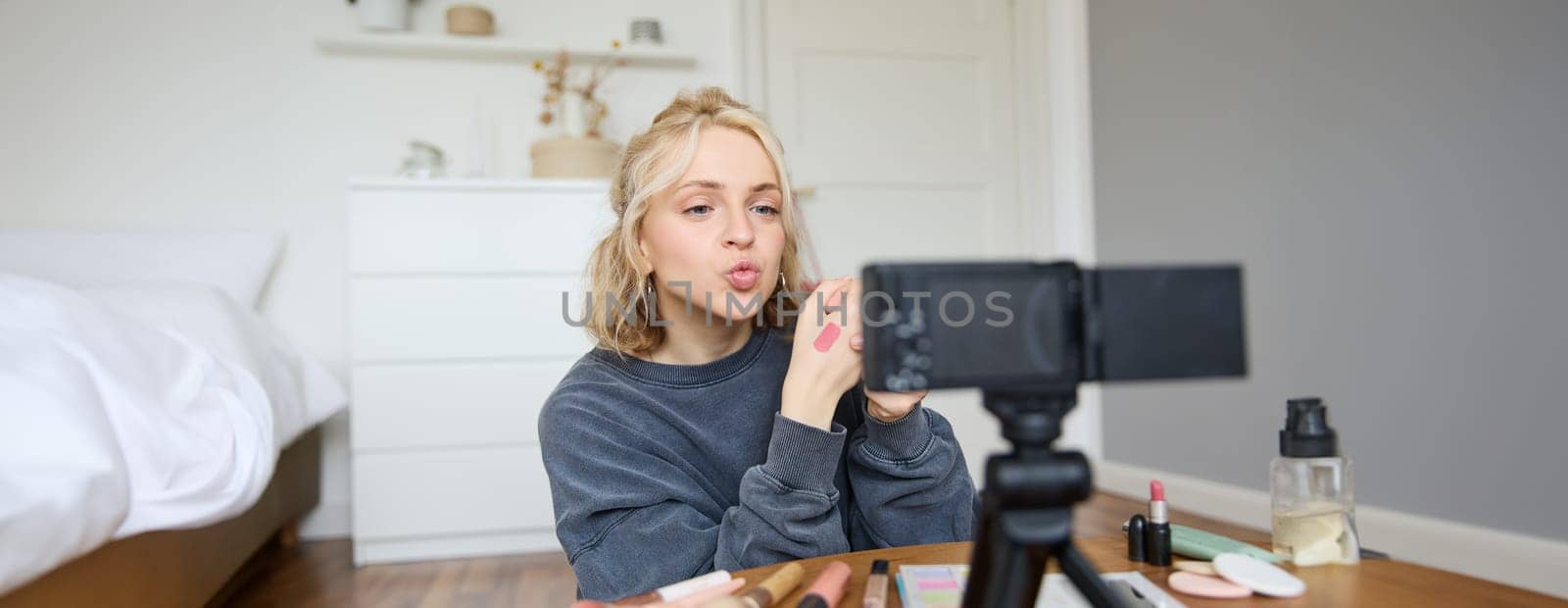 Portrait of young creative social media content creator, woman showing lipstick swatches on her hand, recording video about beauty and makeup, sitting in her room in front of digital camera.