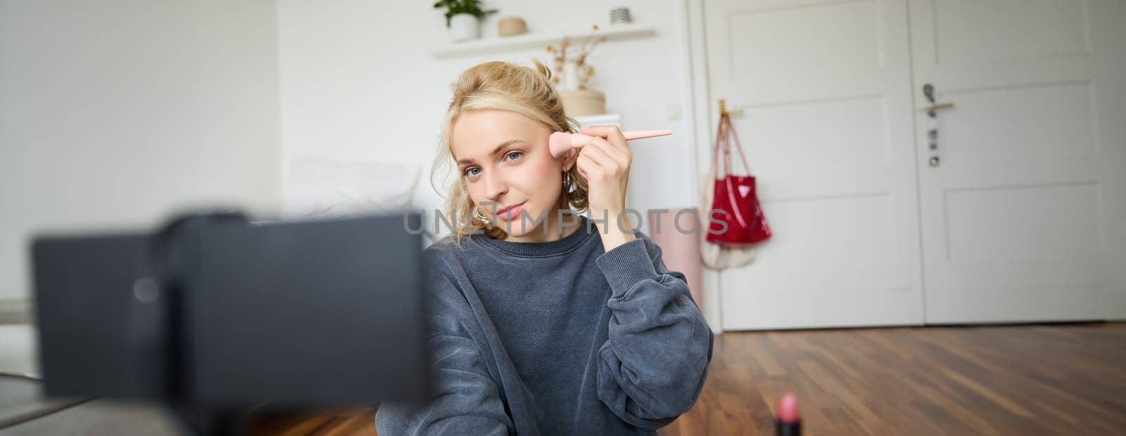 Portrait of young beautiful woman applies makeup blushes with a brush and records herself on digital camera, creates content for beauty blog, social media account by Benzoix