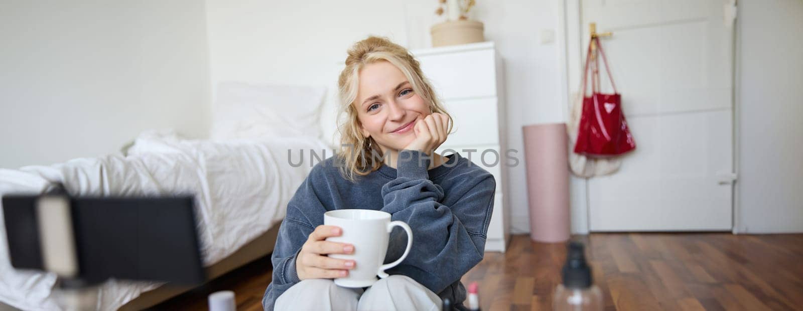 Close up portrait of blond young woman, sitting in her room, looking at camera, holding cup of tea, recording cosy lifestyle blog, vlogger creating content for social media account by Benzoix