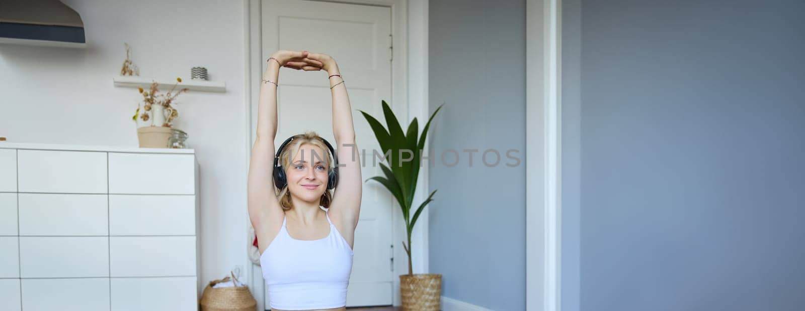 Vertical shot of smiling, active and healthy woman workout at home, follows fitness instructions, watches video on laptop in headphones, stretches her arms.