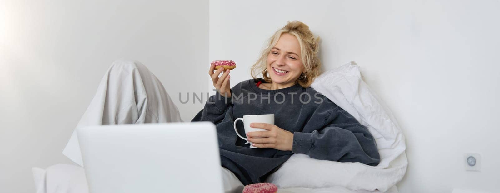 Lifestyle and people concept. Happy blond woman lying in bed with food, drinking tea and eating doughnut, resting at home, spending time alone, watching tv show by Benzoix