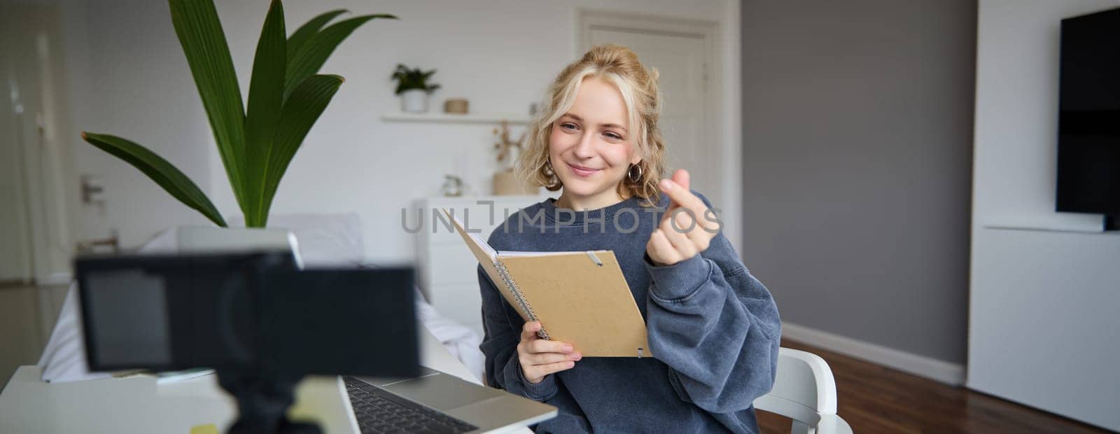 Beautiful young woman in her room, sits indoors, records video on digital camera, holds notebook and her notes, shows heart finger sign.