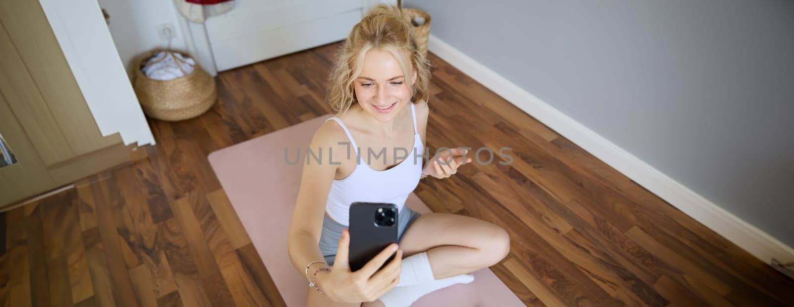 Young sporty fitness trainer, yoga instructor does live stream, takes selfies on smartphone while sitting on rubber mat at home, recording her workout training session.