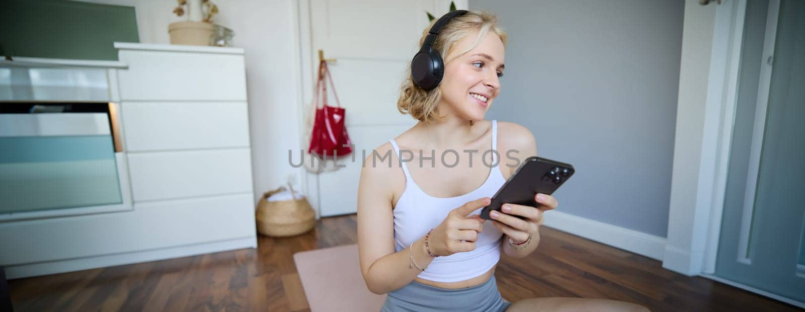 Portrait of smiling, healthy young woman in headphones, holding smartphone, working out at home, using fitness application on mobile phone. Wellbeing and sport concept