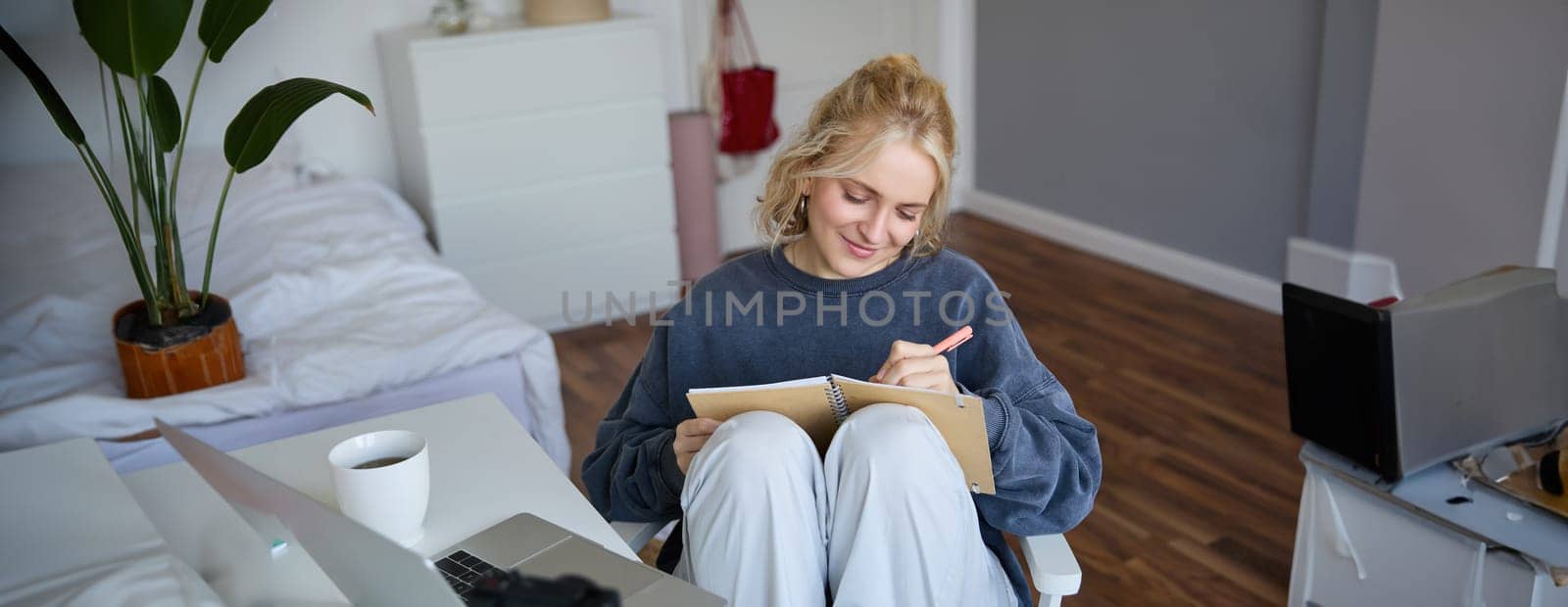 Portrait of young woman writing in diary, making notes in notebook, sitting on chair in a room in front of laptop, student doing homework by Benzoix