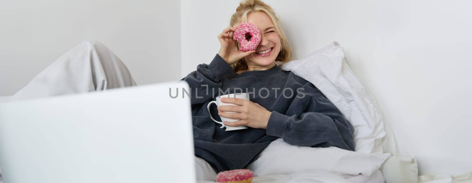 Portrait of happy cute woman, lying in bed with laptop, drinking tea, looking through doughnut hole and smiling at camera.