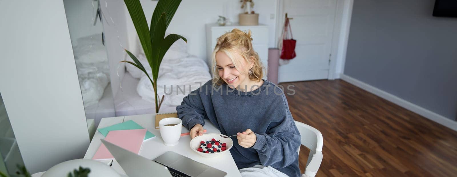 Image of smiling young woman watching videos on laptop while eating breakfast and drinking tea, looking at screen, sitting in her room by Benzoix