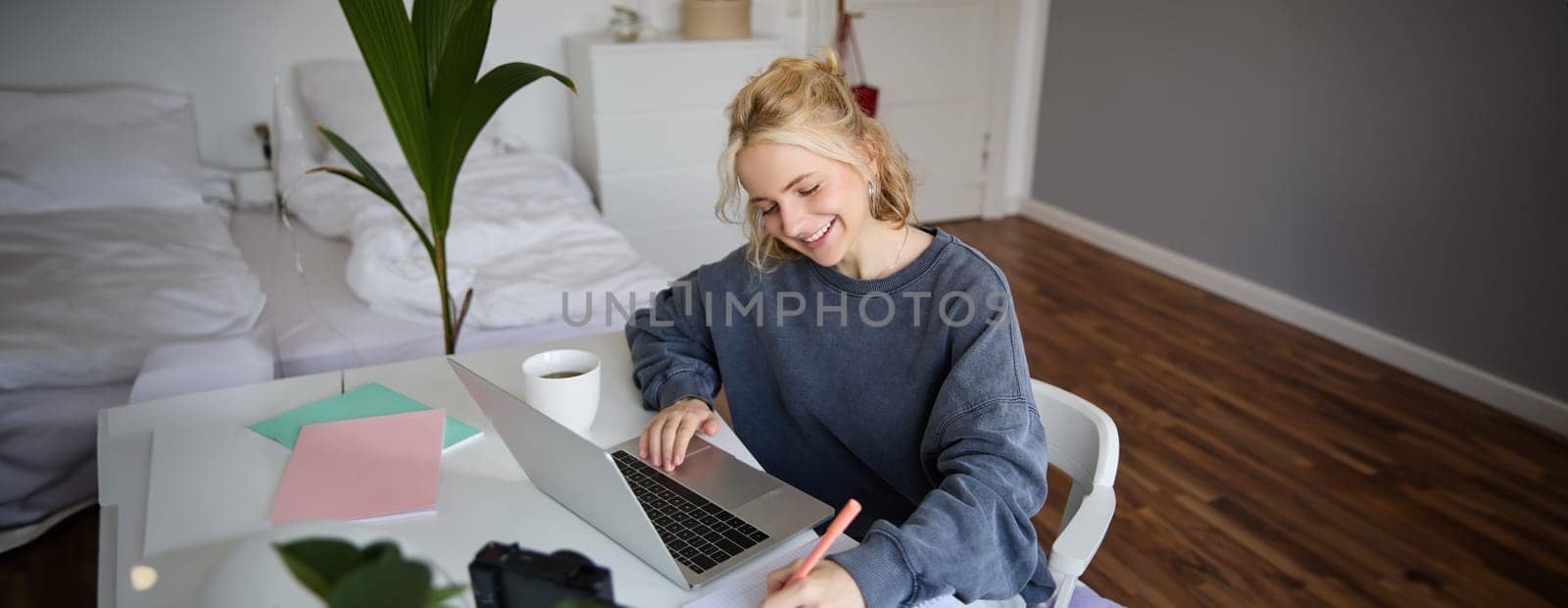 Portrait of smiling, beautiful blond woman, writing down notes, doing homework, studying from home, doing distance learning, online course, working remotely in her room by Benzoix