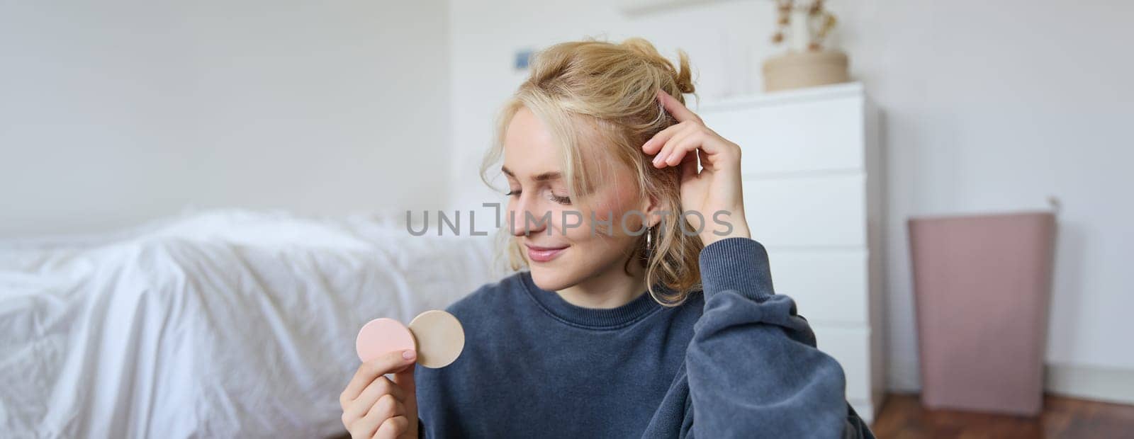 Portrait of young woman chatting on live stream about makeup, sits on floor in bedroom, showing beauty products to followers.