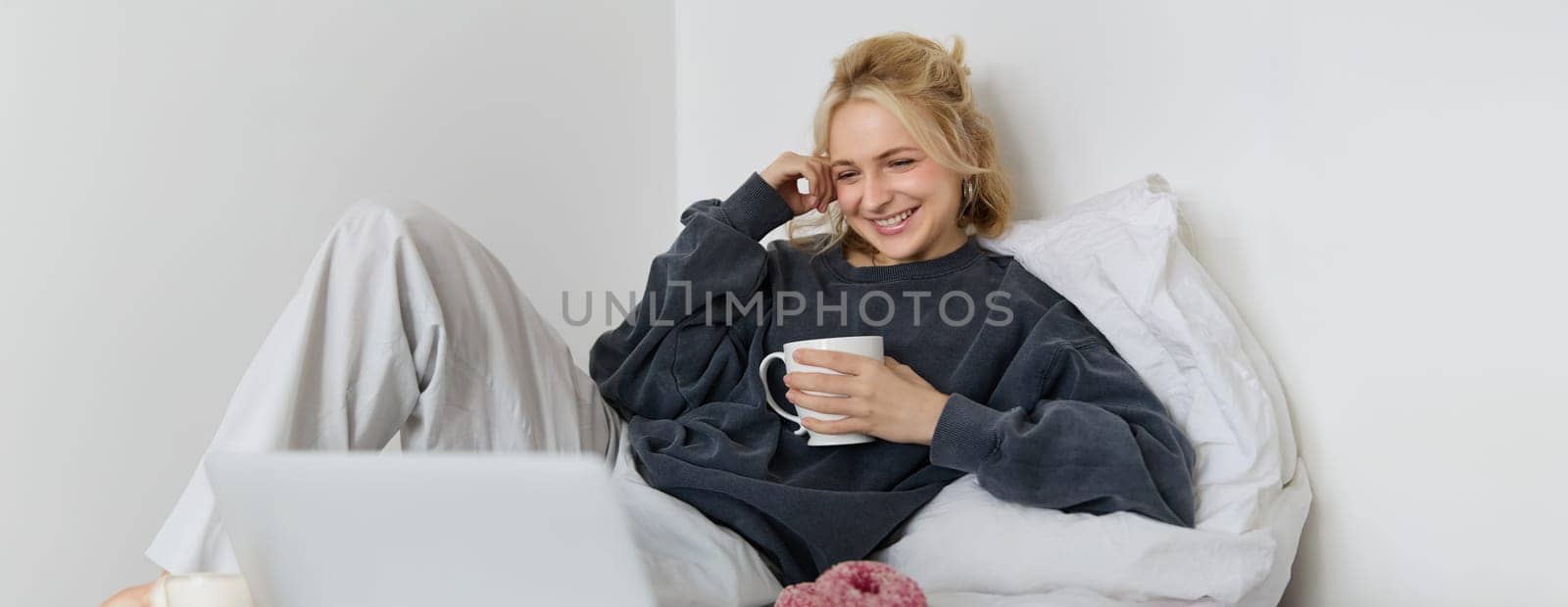 Portrait of candid, happy young woman lying in bed, looking at laptop screen, holding cup of tea and eating doughnut, staying at home on weekend, spending quite time alone by Benzoix