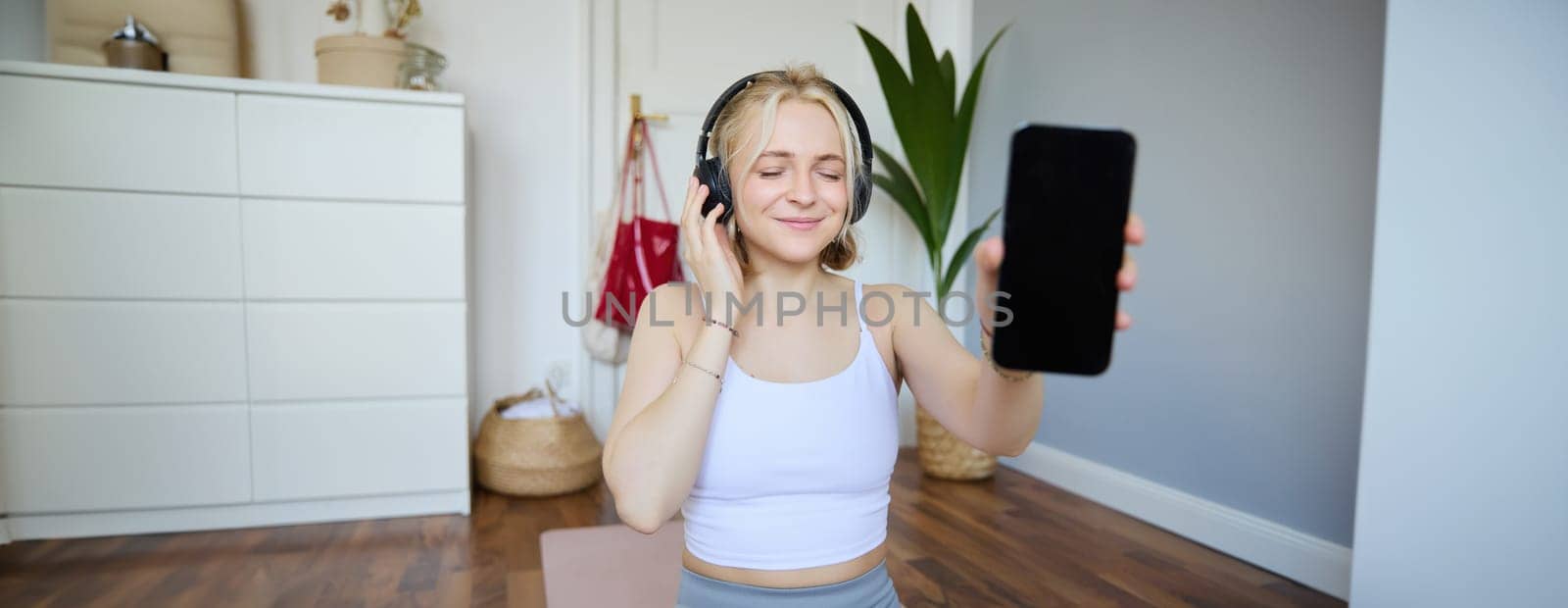 Portrait of smiling, beautiful young woman, showing her smartphone screen, wearing headphones, looking satisfied with sound quality by Benzoix