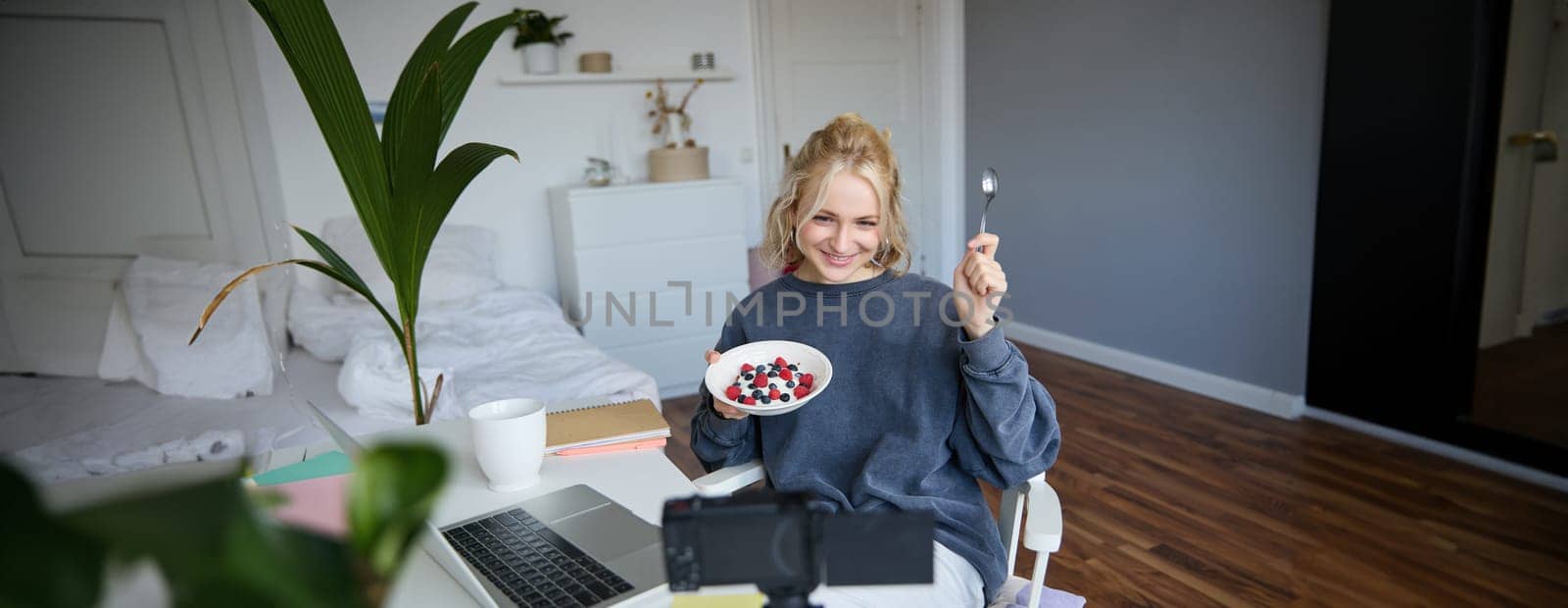 Portrait of cute young girl vlogger, showing her breakfast on camera, recording vlog in her bedroom, holding dessert, eating.