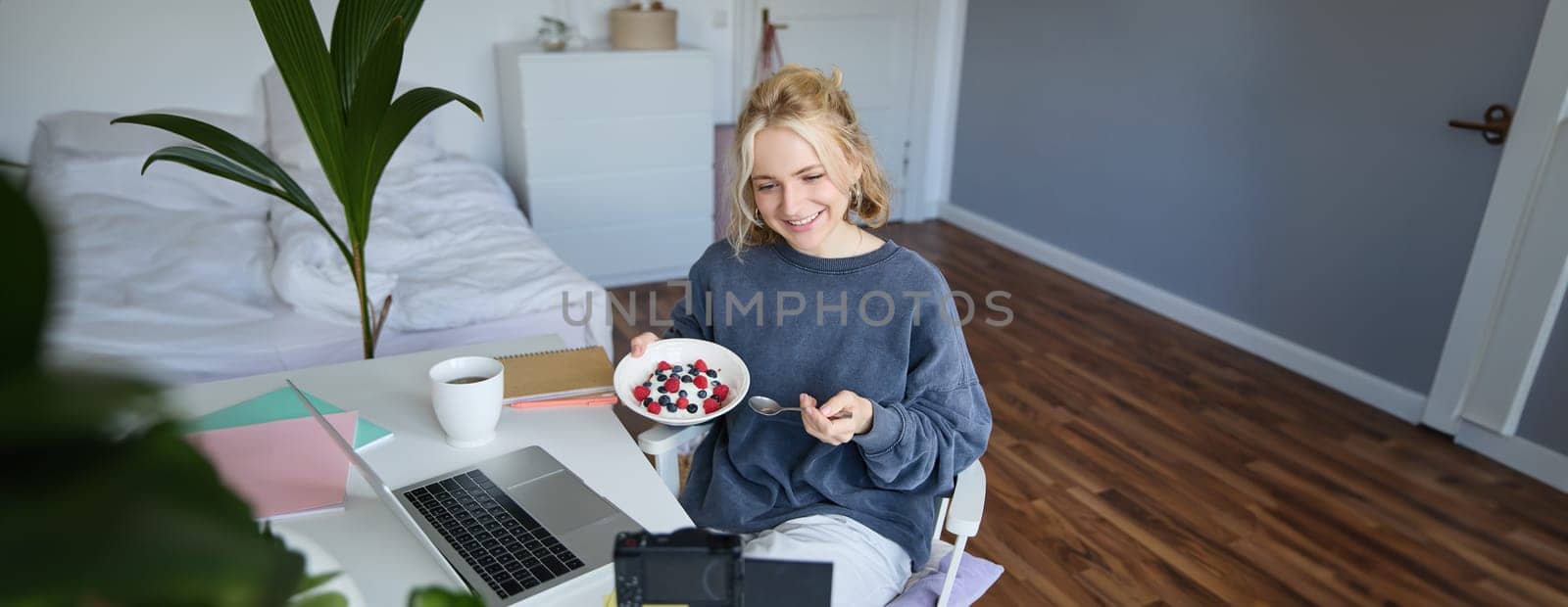 Portrait of smiling young female vlogger, recording and creating content in her room, eating in front of camera, talking, making lifestyle blog.