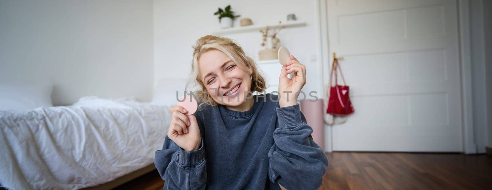 Portrait of young woman, content creator, showing beauty makeup products at camera, sitting on floor in bedroom and smiling.