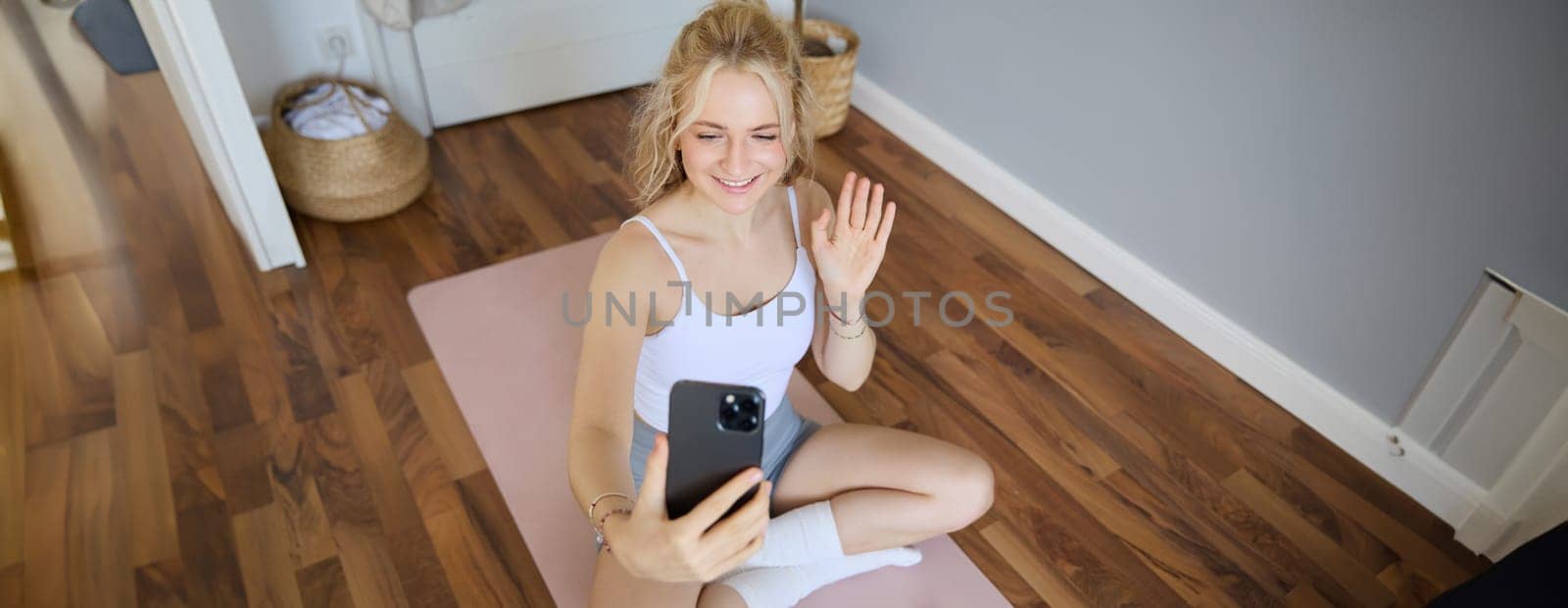 Portrait of fit, sporty young fitness instructor, woman gives online workout sessions to clients, waves hand at smartphone, sits on yoga mat and shows exercises.