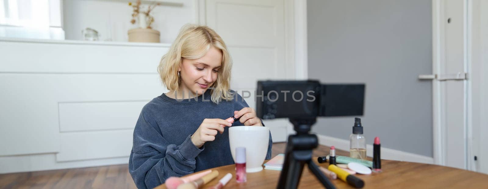 Portrait of young smiling woman in her room, recording video on camera, lifestyle vlog for social media, holding mascara, reviewing her makeup beauty products, showing how to use cosmetics.