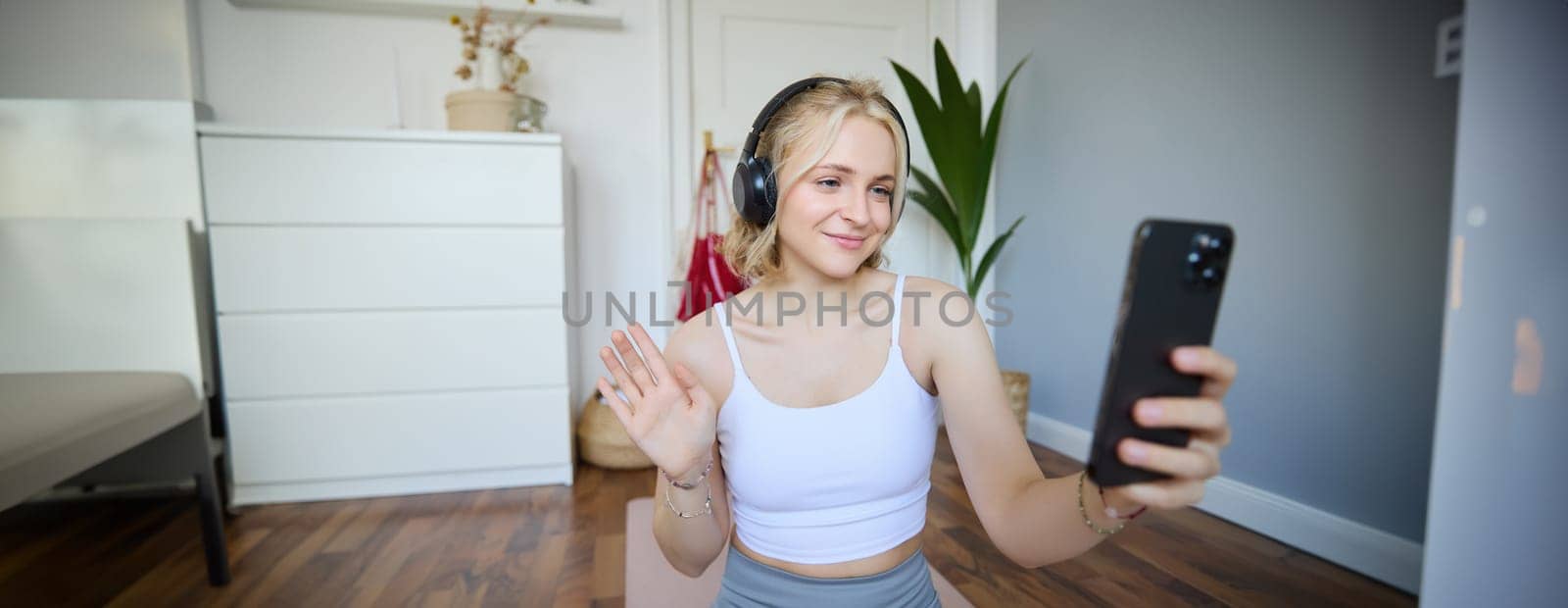 Portrait of young woman, fitness instructor doing online workout session on smartphone, waving hand at camera and saying hello, sitting on rubber mat in a room by Benzoix