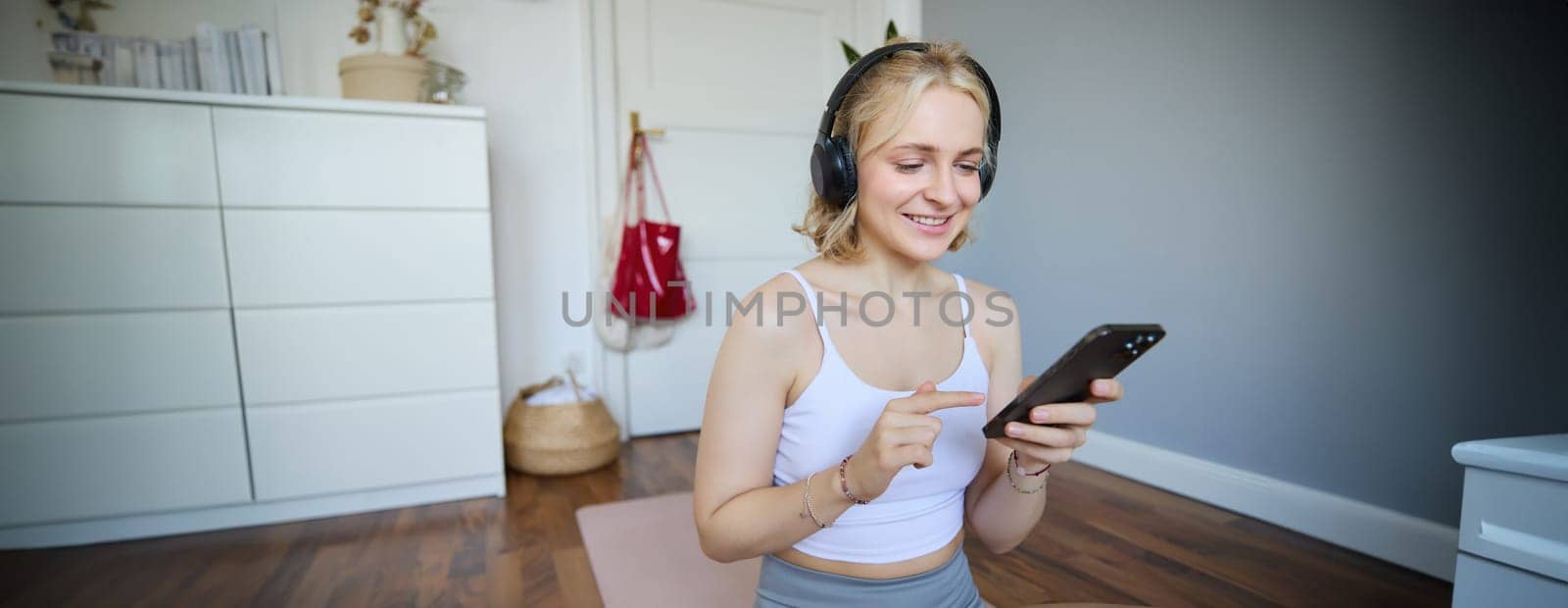 Portrait of young sporty woman in wireless headphones, sitting with smartphone on rubber mat, using workout training app, fitness application on her phone, listening in earphones.