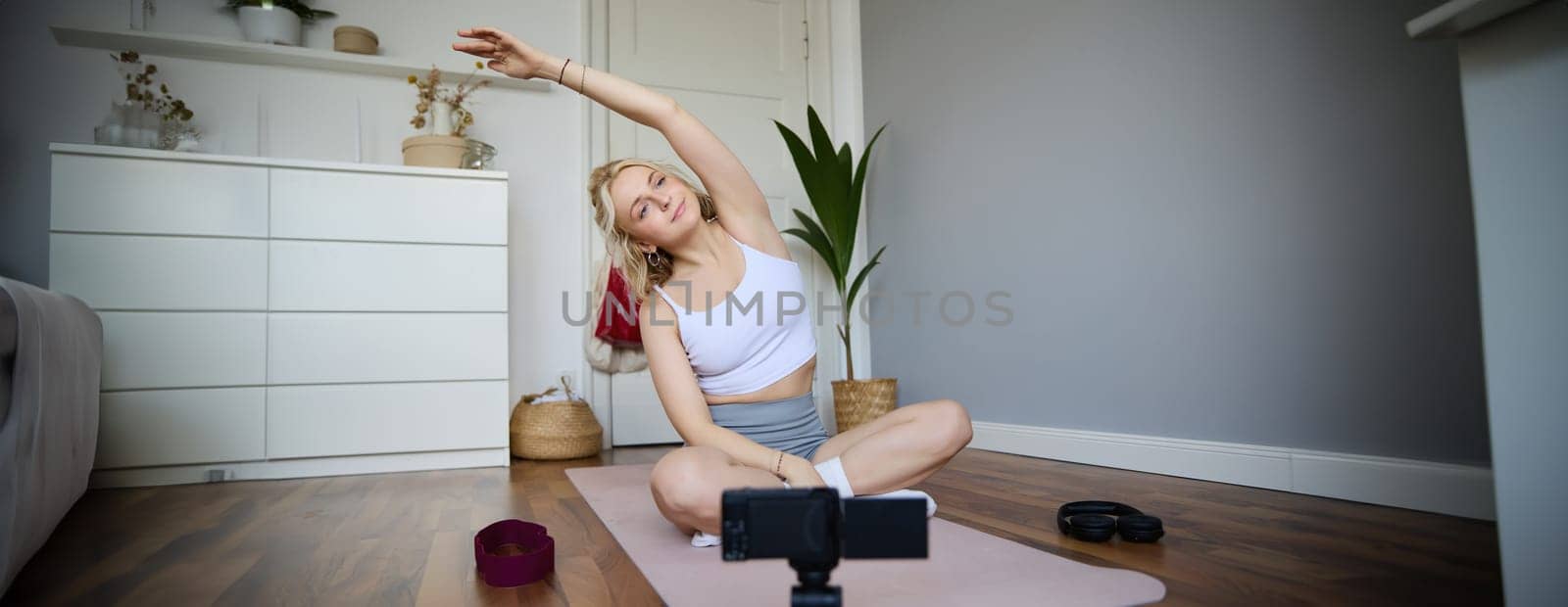 Portrait of young blogger, yoga content creator, showing exercises, recording video of herself working out at home on rubber mat.