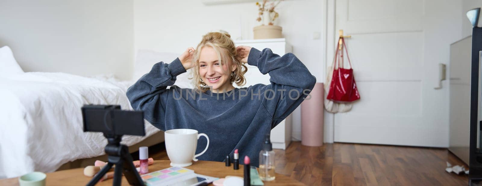 Portrait of cute blond girl, lifestyle blogger sits on floor with video camera and stabiliser, shows how to do hairstyle and makeup, does casual vlogging for social media, sits in room.