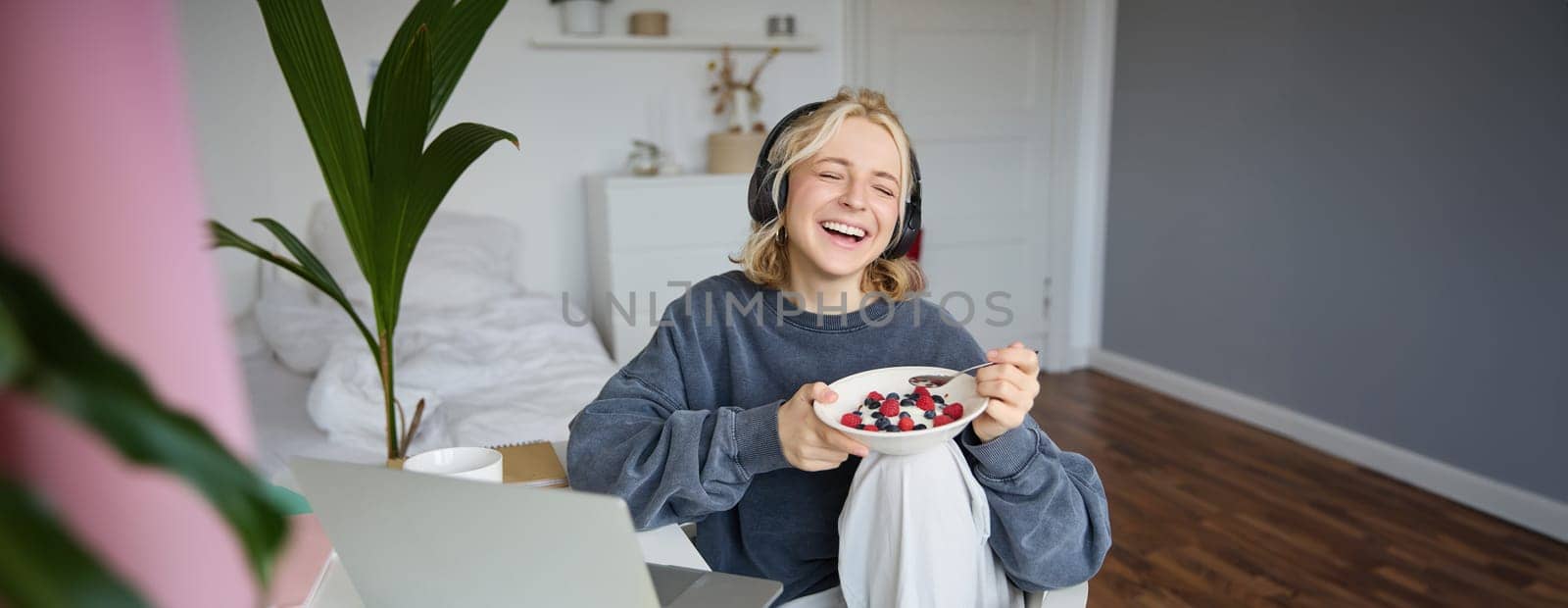 Portrait of happy young blond woman, sitting in a room, watching movie on laptop and eating healthy breakfast, drinking tea, resting on weekend.