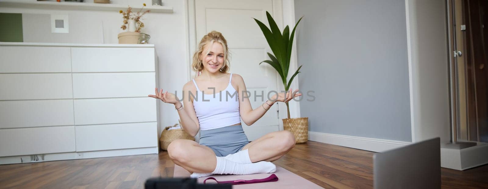 Young female athlete, fitness instructor woman sits on floor rubber mat, recording video on digital camera, showing how to workout, explaining exercises.