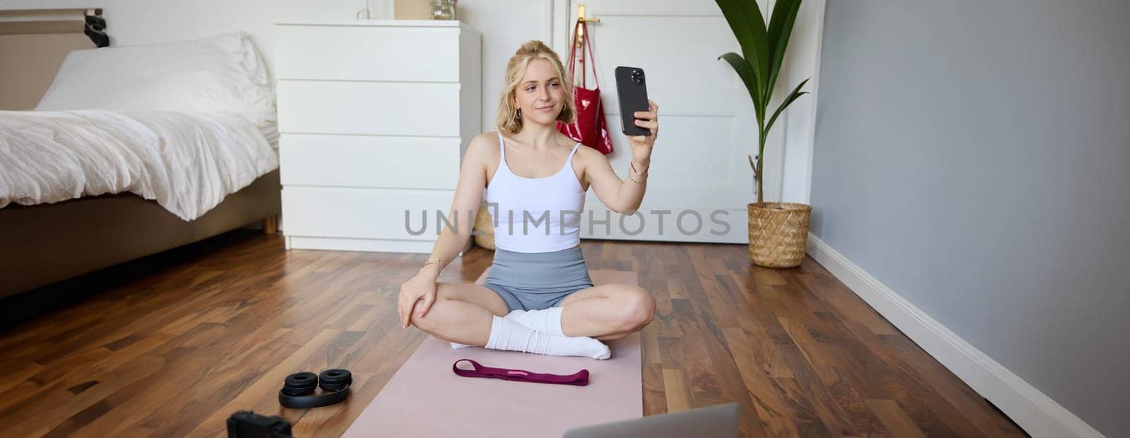 Portrait of beautiful lifestyle blogger, recording herself working out at home, sitting on rubber mat doing yoga in her room, taking selfie on smartphone, using digital video camera by Benzoix