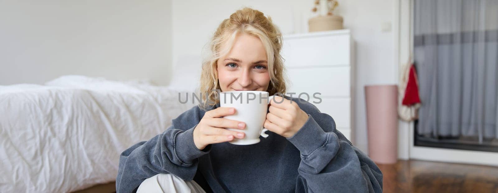 Image of young teenage girl sitting in her bedroom on floor, drinking cup of tea and enjoying day at home, smiling and looking at camera by Benzoix