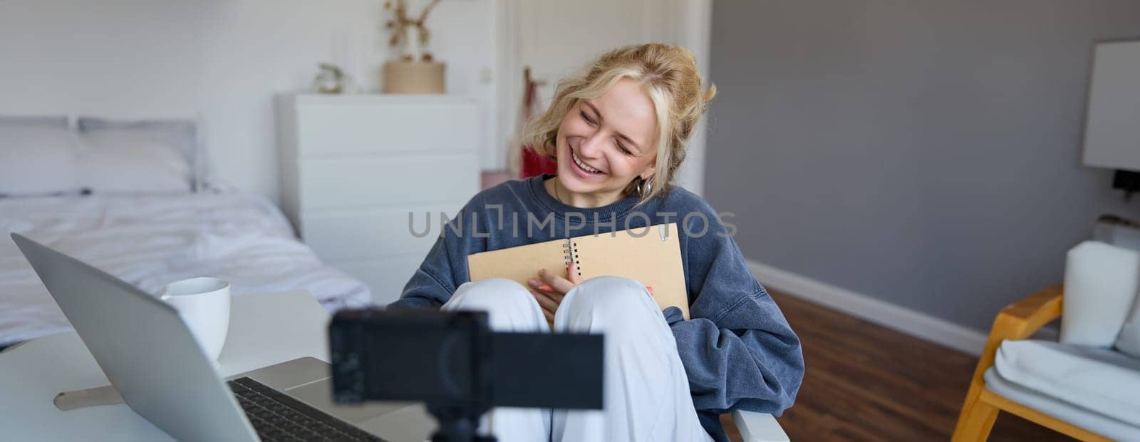 Social media and lifestyle content concept. Young happy smiling woman, sits in her room with notebook, talks at digital camera, having conversation with followers, does lifestyle vlog by Benzoix