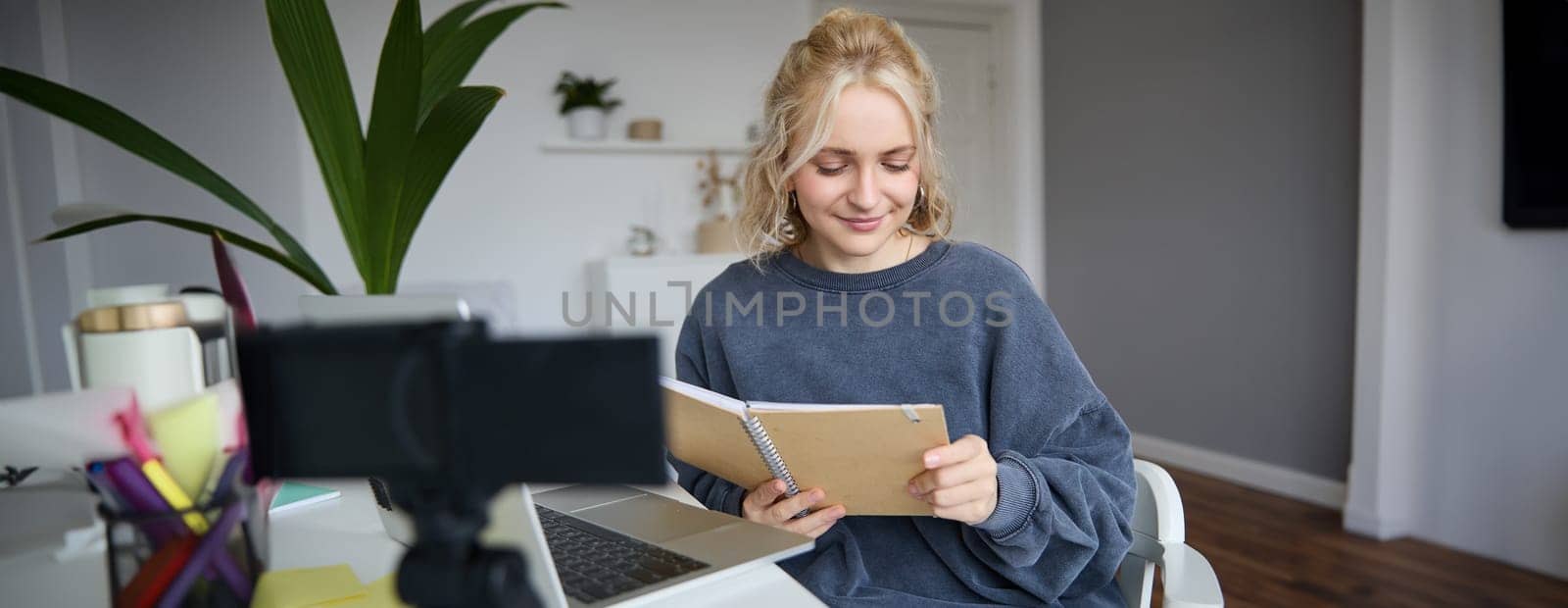 Portrait of young woman with notes, working from home, recording video on digital camera, live streaming from her room, reading from notebook and smiling by Benzoix