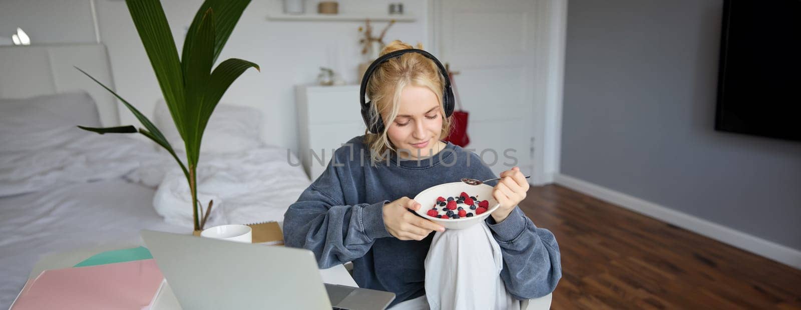 Portrait of smiling young blond woman in headphones, sitting in room, watching movie on laptop, eating breakfast and drinking tea, having lunch in front of computer screen by Benzoix