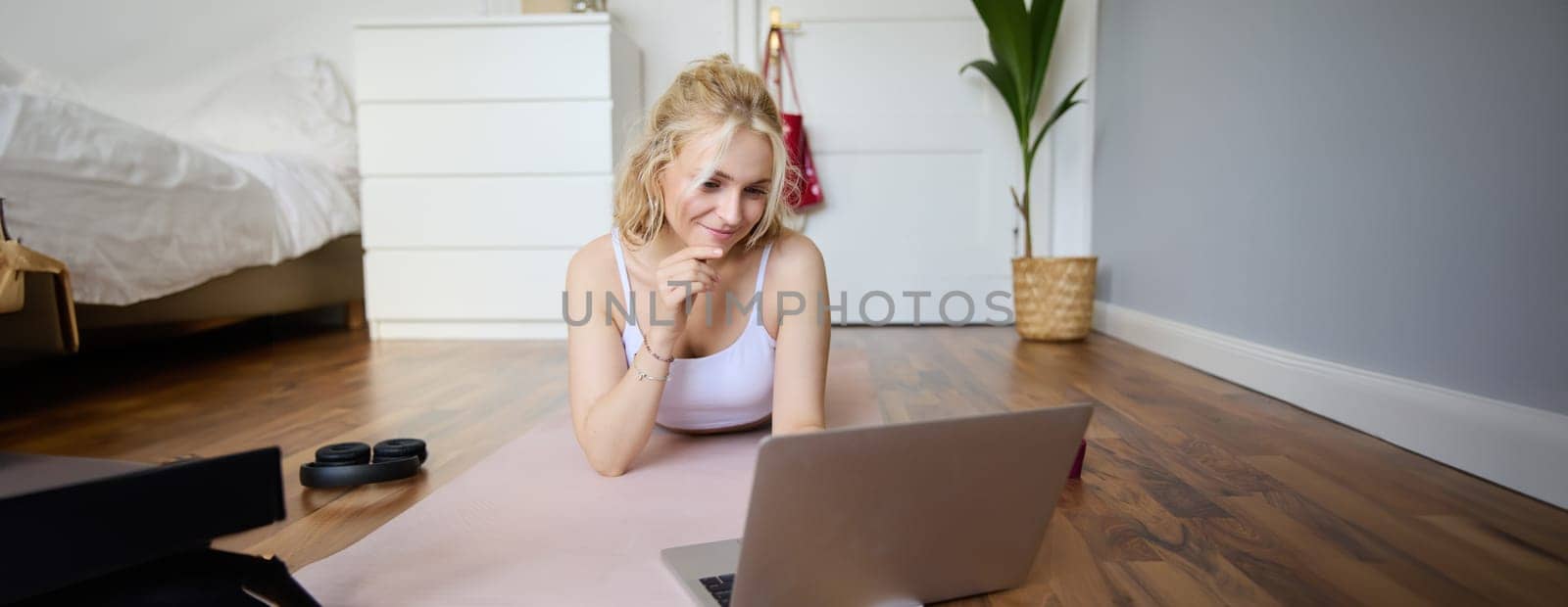 Portrait of beautiful blond woman looking at fitness video tutorials on laptop, lying on rubber yoga mat, following workout instructions online.