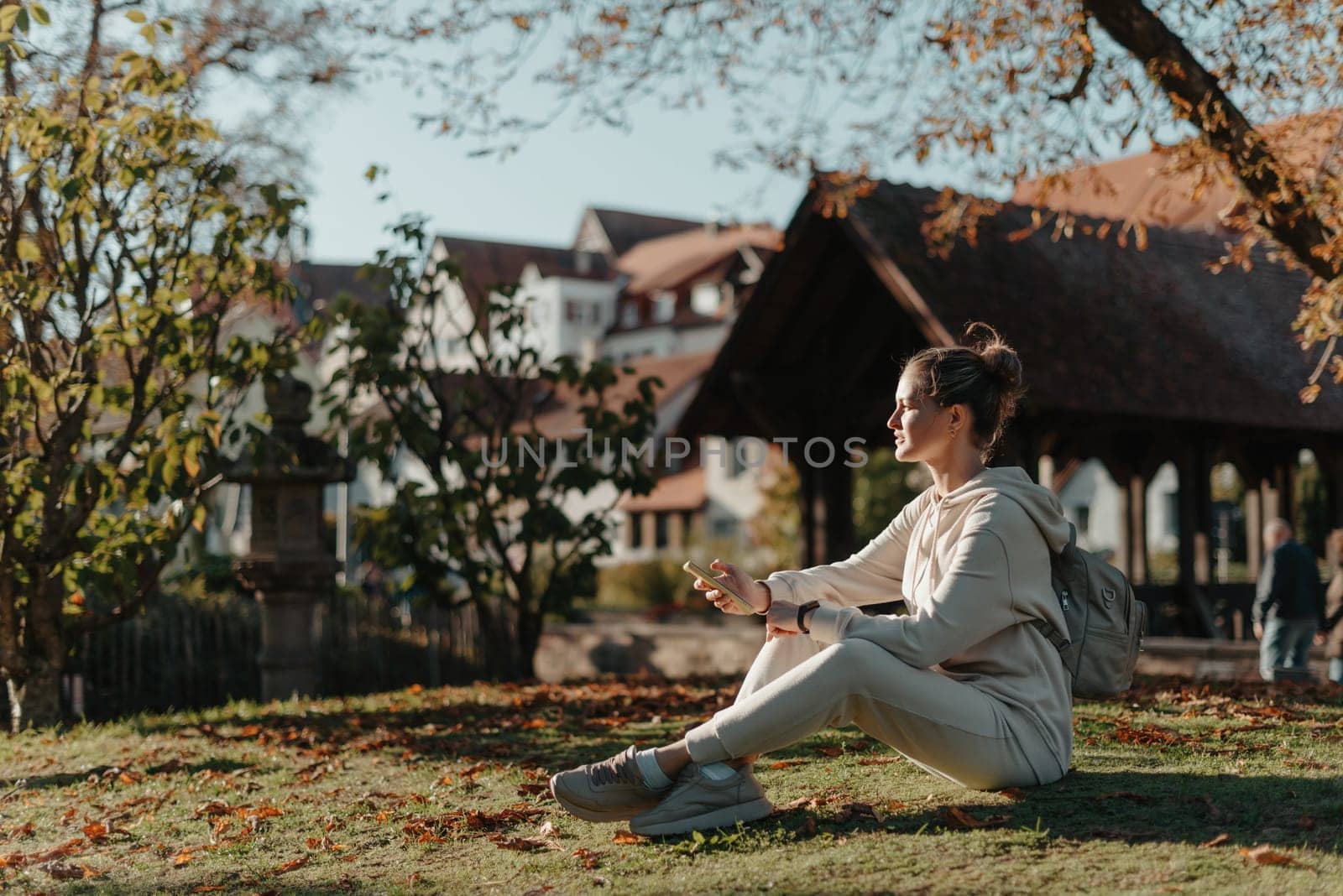 Young fashionable teenage girl with smartphone in park in autumn sitting at smiling. Trendy young woman in fall in park texting. Retouched, vibrant colors. Beautiful blonde teenage girl wearing casual modern autumn outfit sitting in park in autumn. Retouched, vibrant colors, brownish tones.