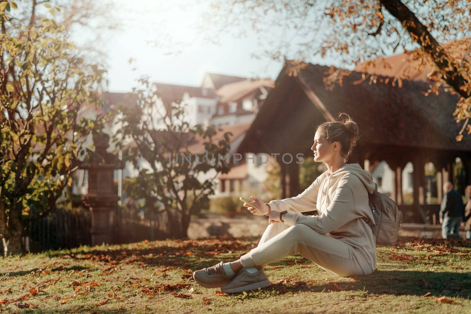 Young fashionable teenage girl with smartphone in park in autumn sitting at smiling. Trendy young woman in fall in park texting. Retouched, vibrant colors. Beautiful blonde teenage girl wearing casual modern autumn outfit sitting in park in autumn. Retouched, vibrant colors, brownish tones.