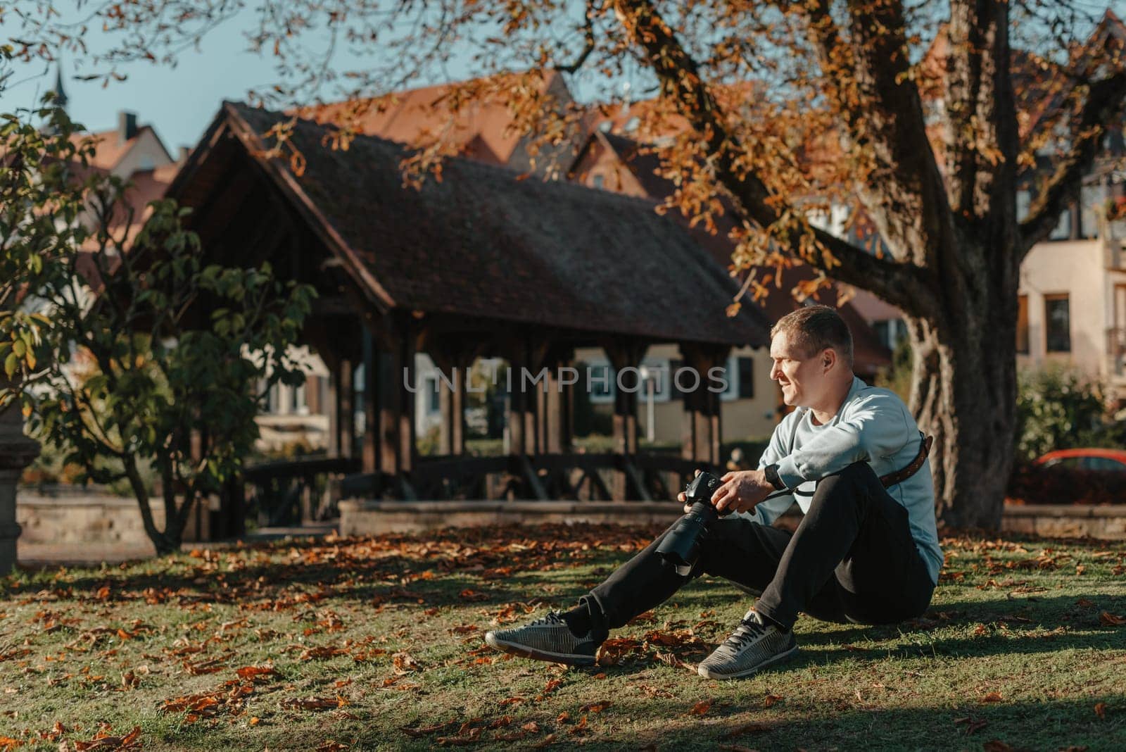 Professional Photographer Taking Picture Of Beautiful Autumn Park. Man Professional Photographer Sit With Camera And With Smartphone In Autumn Park. Retouched, Vibrant Colors, Brownish Tones. by Andrii_Ko