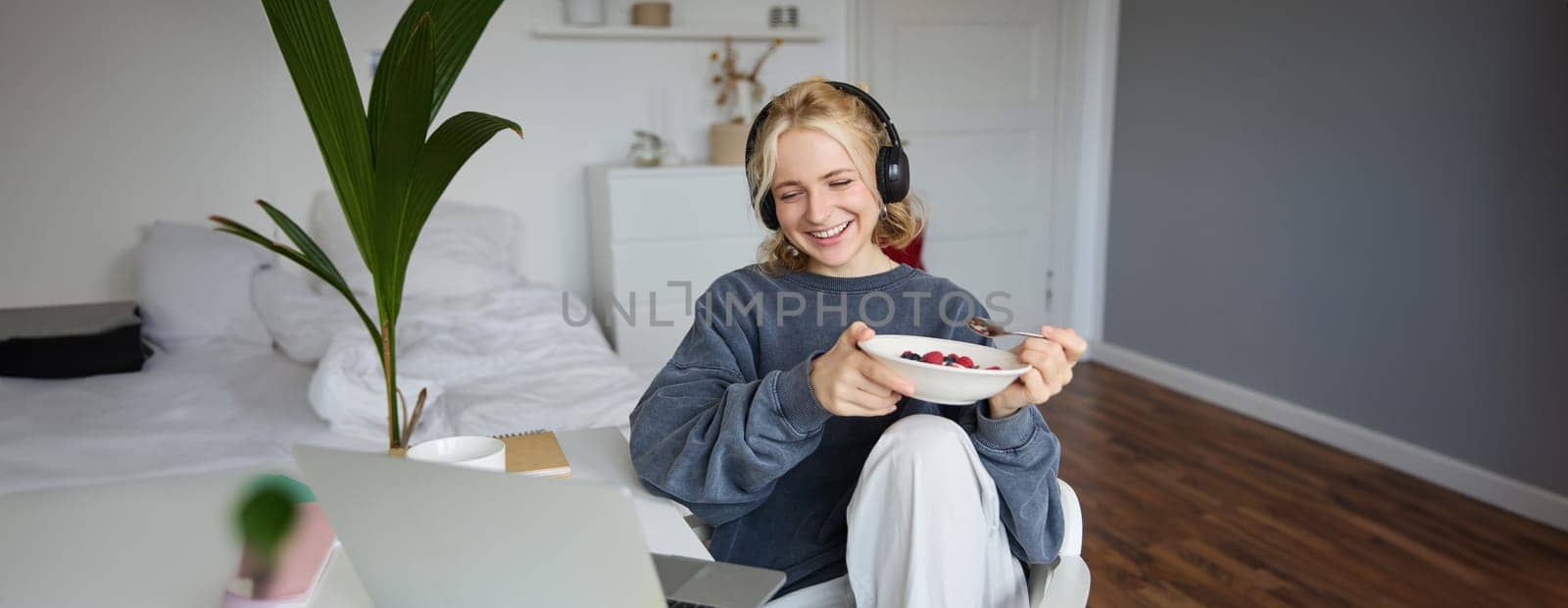 Portrait of smiling young woman, watching tv show in headphones, eating breakfast and looking at laptop screen.