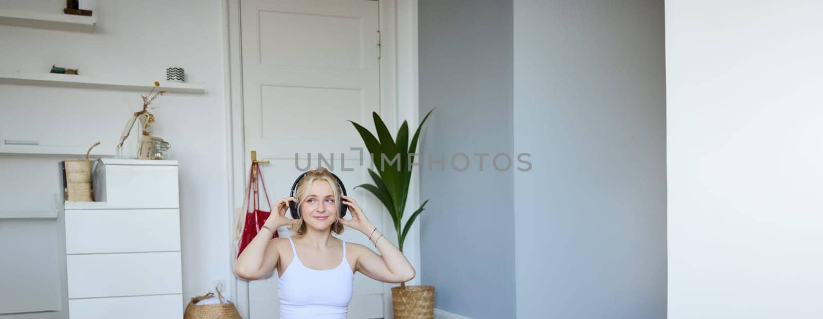 Vertical shot of smiling young woman using video tutorials for workout at home, sitting with laptop in wireless headphones on yoga mat, follows fitness instructor on social media, repeats exercises.