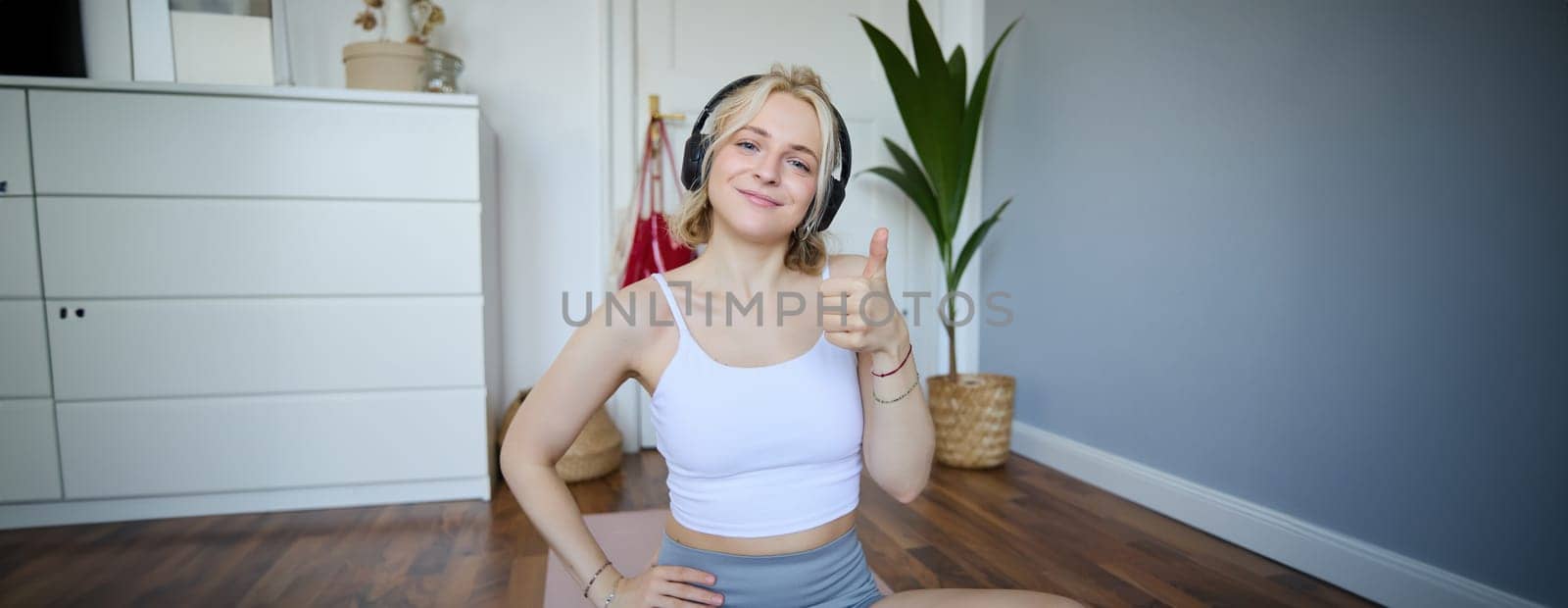 Portrait of beautiful, healthy woman in headphones, showing thumbs up, sitting on rubber mat, recommending yoga online training video, pleased with exercises for home training.