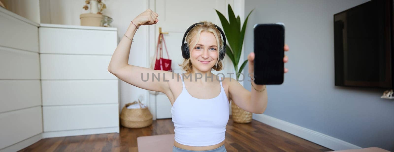 Portrait of beautiful and fit young woman, staying healthy working out at home, showing mobile app, blank screen, flexing biceps, recommending smartphone application for exercises.
